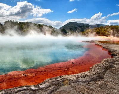 Vibrant geothermal pool with orange and blue waters emits steam under a bright blue sky. Lush forest and distant mountains provide a serene backdrop.