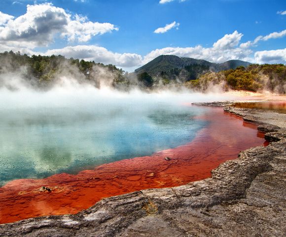 Vibrant geothermal pool with orange and blue waters emits steam under a bright blue sky. Lush forest and distant mountains provide a serene backdrop.