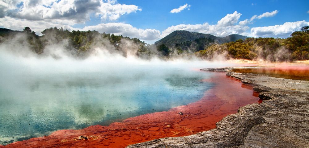 Vibrant geothermal pool with orange and blue waters emits steam under a bright blue sky. Lush forest and distant mountains provide a serene backdrop.