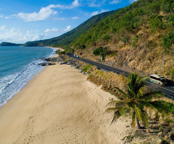 Aerial view of a serene coastal road flanked by lush green hills and a sandy beach, with gentle waves lapping the shore under a blue sky.