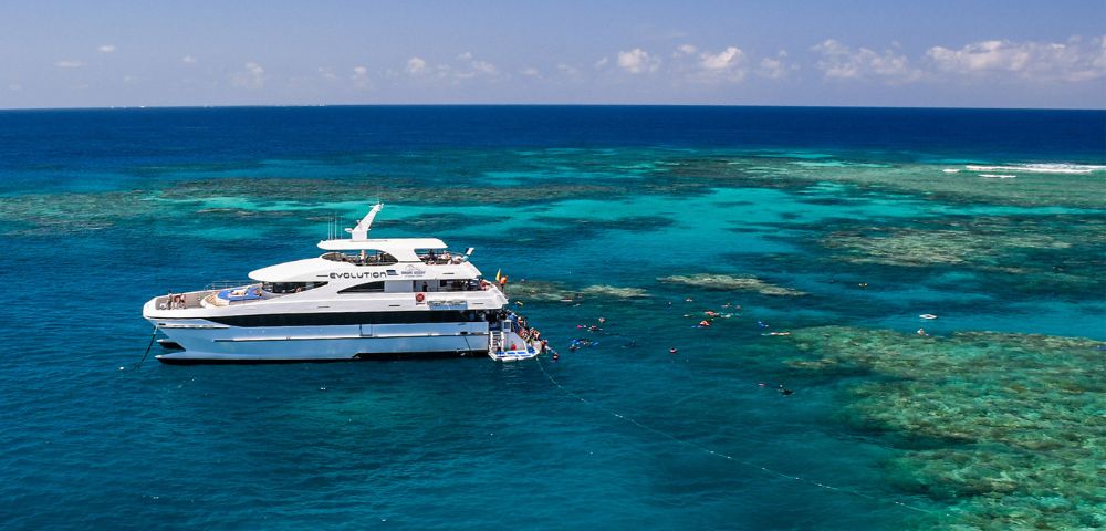 A luxury yacht anchored in clear turquoise waters near a vibrant coral reef, with snorkelers exploring the marine life. The scene is calm and sunny.