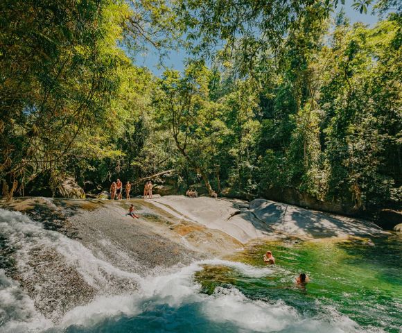 People enjoy a sunny day at a natural rock slide surrounded by lush green forest. Some swim in the clear pool, while others stand on the rocks.