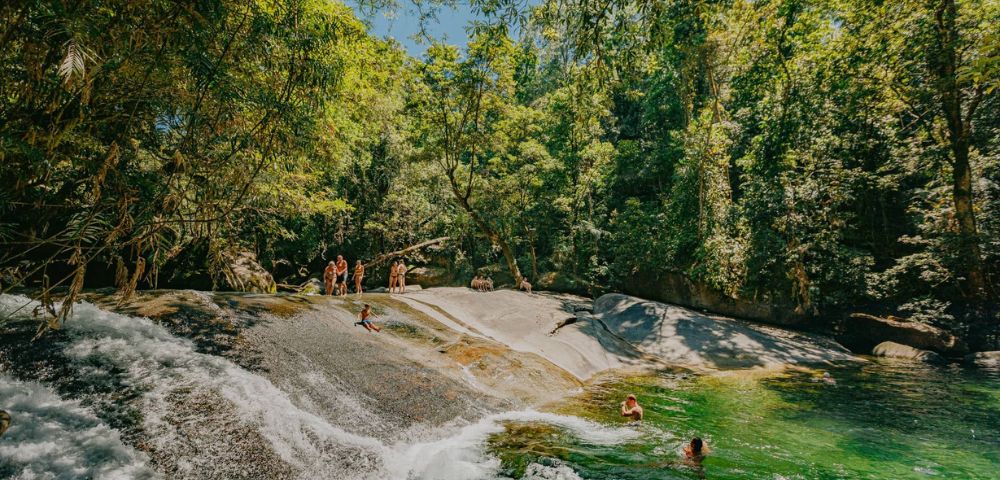 People enjoy a sunny day at a natural rock slide surrounded by lush green forest. Some swim in the clear pool, while others stand on the rocks.