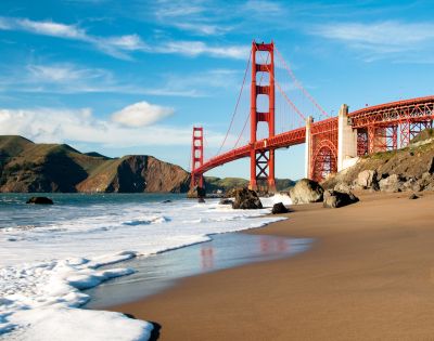 A scenic view of the Golden Gate Bridge under a clear blue sky, with waves gently lapping against a sandy beach and rocky shoreline in the foreground.