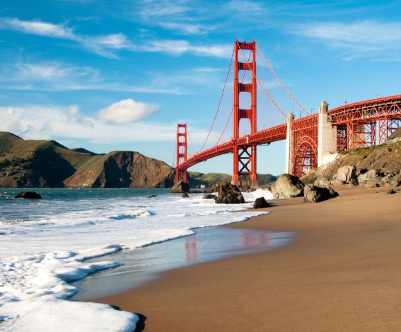 A scenic view of the Golden Gate Bridge under a clear blue sky, with waves gently lapping against a sandy beach and rocky shoreline in the foreground.