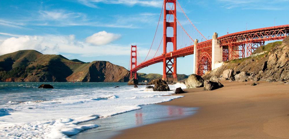 A scenic view of the Golden Gate Bridge under a clear blue sky, with waves gently lapping against a sandy beach and rocky shoreline in the foreground.