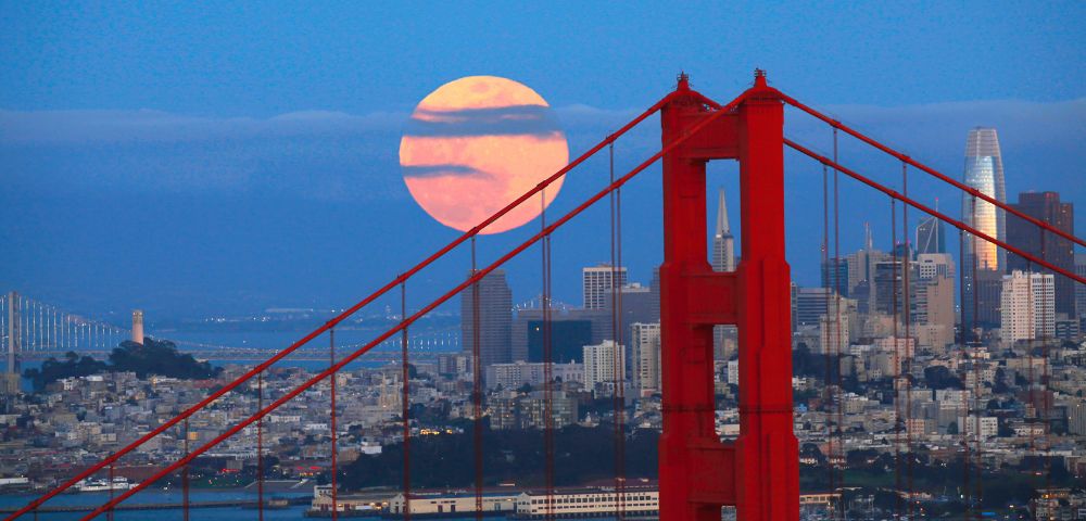 A vivid image of the San Francisco skyline, highlighted by a large full moon rising behind it. The red Golden Gate Bridge stands in the foreground.