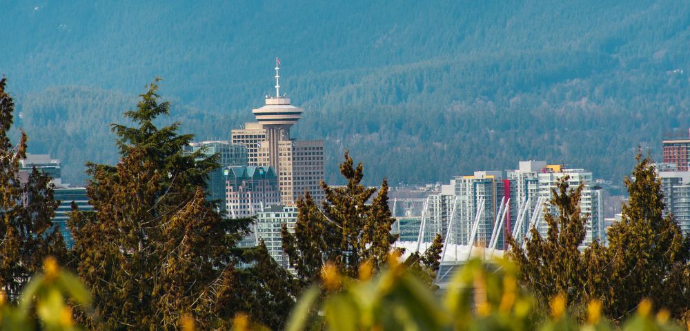 Skyline of a city with a prominent round tower amidst modern buildings, set against a backdrop of lush, forested hills and large trees in the foreground.