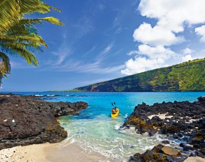 A kayaker paddles in clear turquoise water surrounded by black volcanic rocks and lush green cliffs under a bright blue sky with palm trees nearby.