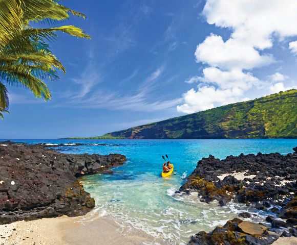A kayaker paddles in clear turquoise water surrounded by black volcanic rocks and lush green cliffs under a bright blue sky with palm trees nearby.
