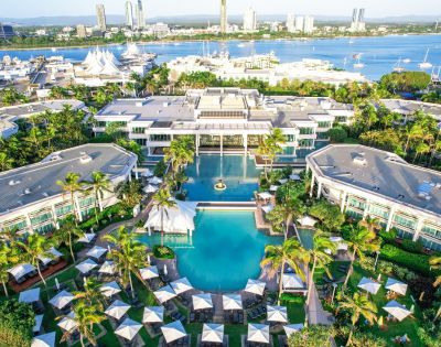 Aerial view of a luxury resort with large pools, surrounded by lush palm trees. Umbrellas dot the landscape, and a marina with boats lies beyond.