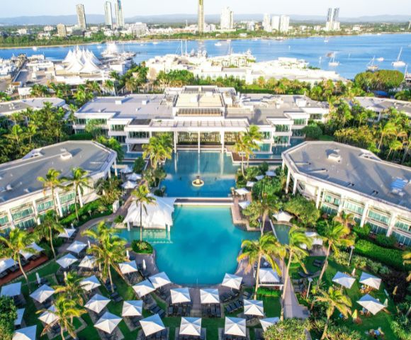Aerial view of a luxury resort with large pools, surrounded by lush palm trees. Umbrellas dot the landscape, and a marina with boats lies beyond.