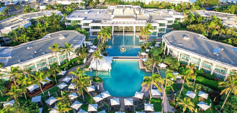 Aerial view of a luxury resort with large pools, surrounded by lush palm trees. Umbrellas dot the landscape, and a marina with boats lies beyond.