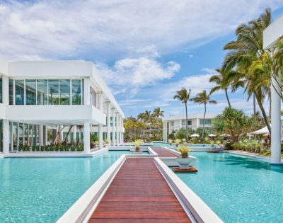 Modern resort with sleek white buildings surrounded by a large, serene pool and wooden walkways. Palm trees and blue skies create a tropical ambiance.