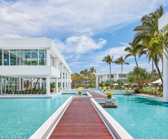 Modern resort with sleek white buildings surrounded by a large, serene pool and wooden walkways. Palm trees and blue skies create a tropical ambiance.