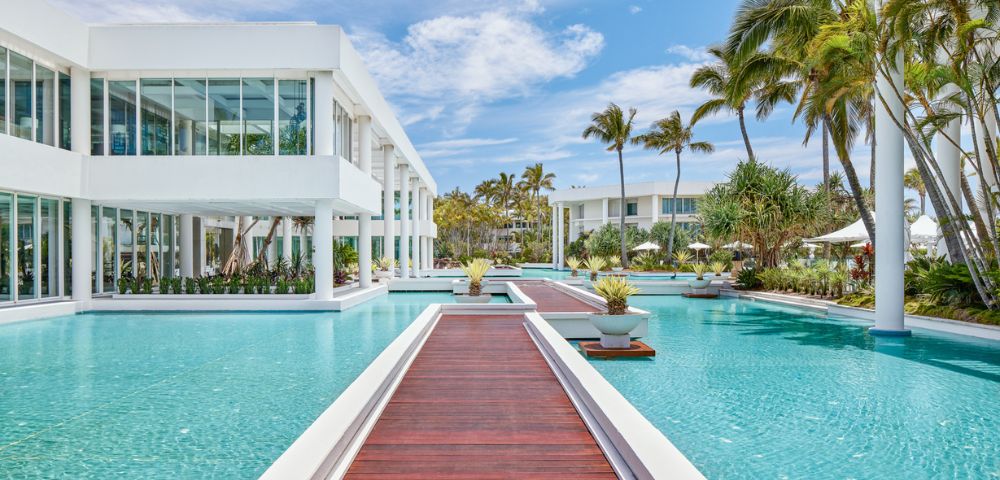Modern resort with sleek white buildings surrounded by a large, serene pool and wooden walkways. Palm trees and blue skies create a tropical ambiance.