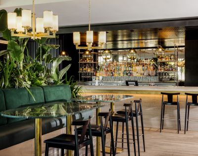 Elegant bar interior with a glass-topped table, dark chairs, and lush plants. Warm lighting and a mirrored back bar create a sophisticated atmosphere.