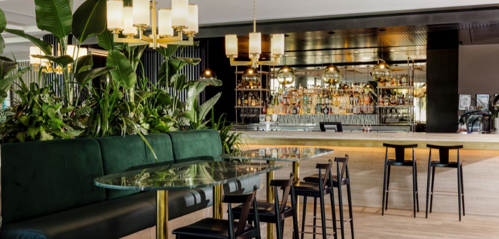 Elegant bar interior with a glass-topped table, dark chairs, and lush plants. Warm lighting and a mirrored back bar create a sophisticated atmosphere.