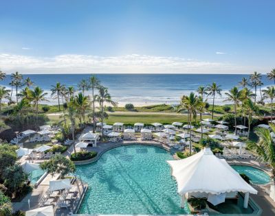 Oceanfront resort with palm trees, sunlit pool, and white canopies flanked by lounge chairs. Serene beach and clear blue sky in the background.