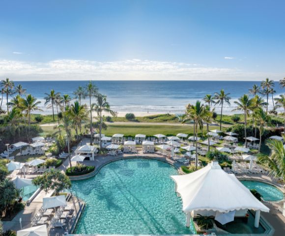 Oceanfront resort with palm trees, sunlit pool, and white canopies flanked by lounge chairs. Serene beach and clear blue sky in the background.