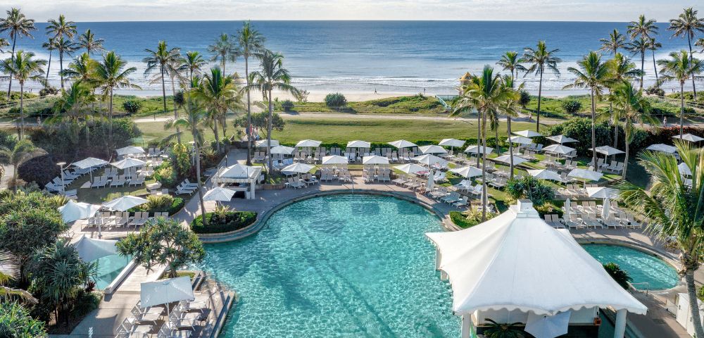 Oceanfront resort with palm trees, sunlit pool, and white canopies flanked by lounge chairs. Serene beach and clear blue sky in the background.