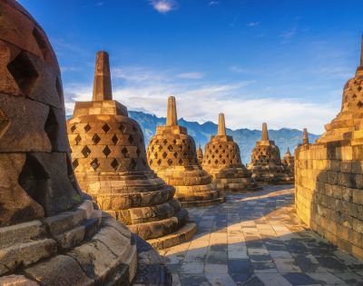 Ancient stone stupas at Borobudur Temple, Indonesia, under a clear blue sky with mountains in the background. The scene is peaceful and historic.