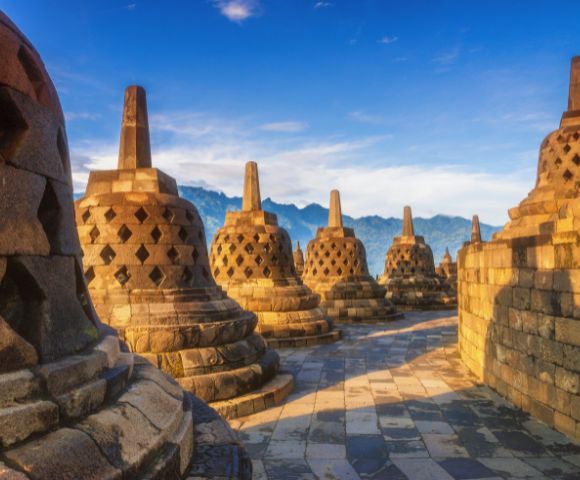 Ancient stone stupas at Borobudur Temple, Indonesia, under a clear blue sky with mountains in the background. The scene is peaceful and historic.