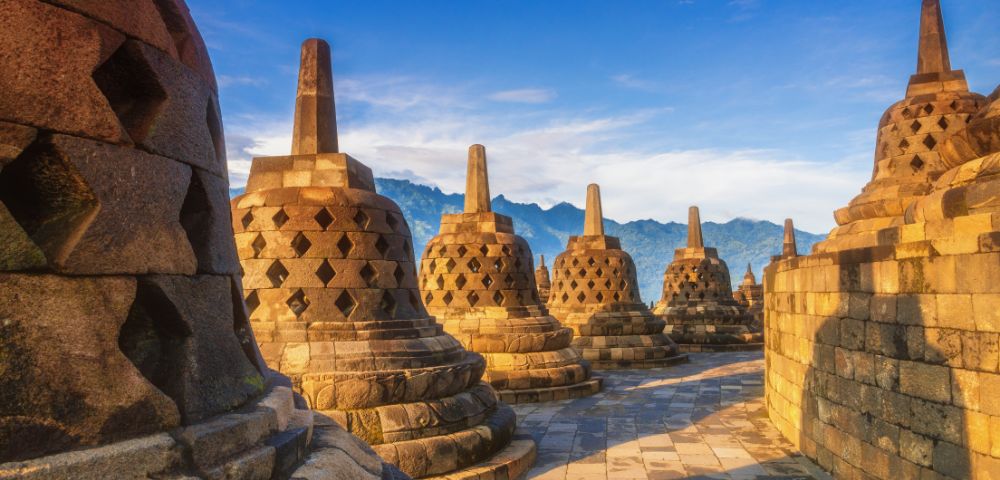 Ancient stone stupas at Borobudur Temple, Indonesia, under a clear blue sky with mountains in the background. The scene is peaceful and historic.
