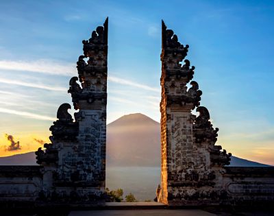 Ornate stone gates frame a distant volcano at sunrise in Bali, Indonesia. The scene conveys serenity, with soft light and a clear sky enhancing the peaceful mood.