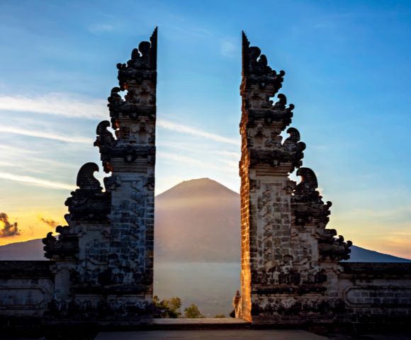 Ornate stone gates frame a distant volcano at sunrise in Bali, Indonesia. The scene conveys serenity, with soft light and a clear sky enhancing the peaceful mood.