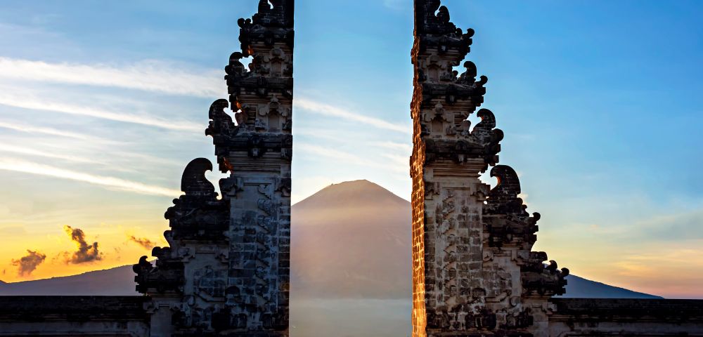Ornate stone gates frame a distant volcano at sunrise in Bali, Indonesia. The scene conveys serenity, with soft light and a clear sky enhancing the peaceful mood.