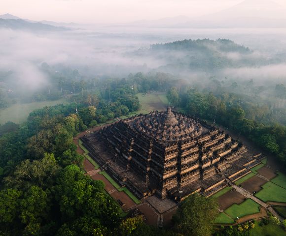 Aerial view of Borobudur Temple in Indonesia, surrounded by lush green trees and morning mist. The ancient, tiered structure exudes a serene ambiance.