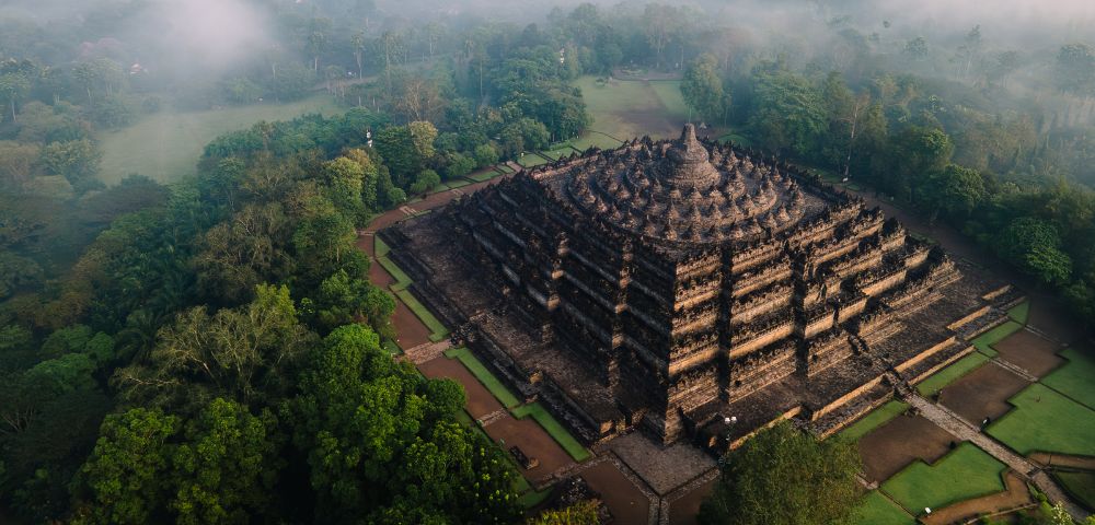 Aerial view of Borobudur Temple in Indonesia, surrounded by lush green trees and morning mist. The ancient, tiered structure exudes a serene ambiance.