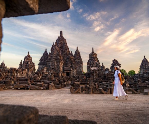 A person in white attire walks past ancient, intricately carved stone temples at sunset, with a sky painted in warm hues, conveying a sense of serenity and history.
