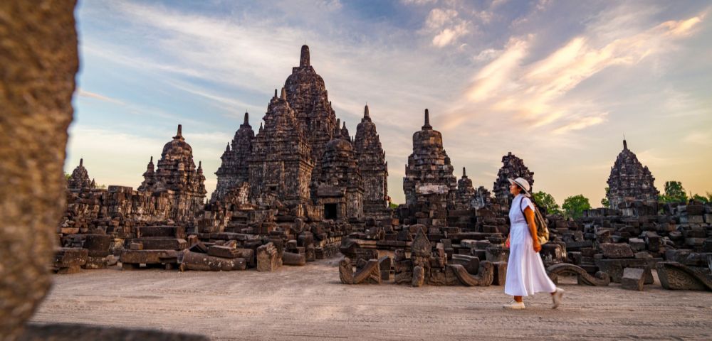 A person in white attire walks past ancient, intricately carved stone temples at sunset, with a sky painted in warm hues, conveying a sense of serenity and history.