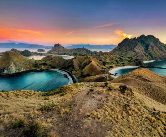 A breathtaking view of Padar Island at sunset, with rolling hills and turquoise bays. The sky is painted with warm hues of orange and pink, creating a serene atmosphere.