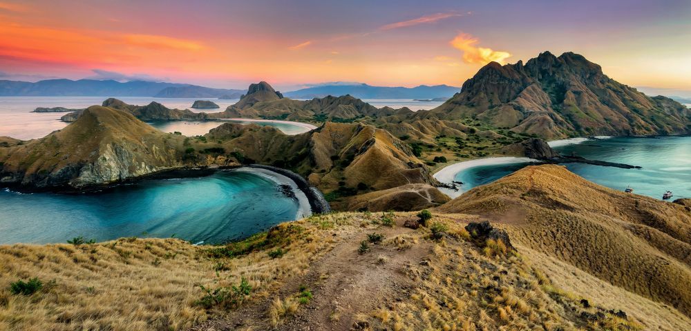 A breathtaking view of Padar Island at sunset, with rolling hills and turquoise bays. The sky is painted with warm hues of orange and pink, creating a serene atmosphere.
