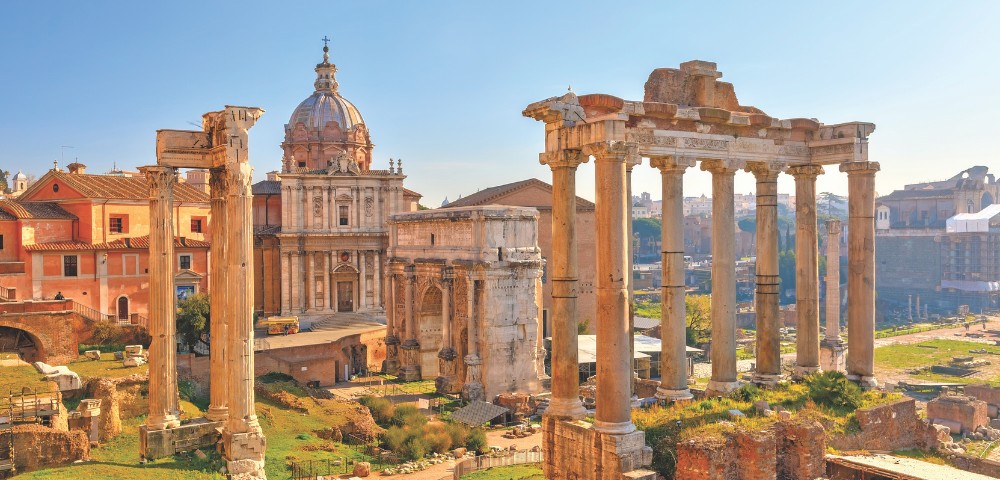 Ancient Roman Forum ruins with tall stone columns, classical architecture, and a domed church in the background under a clear sky.