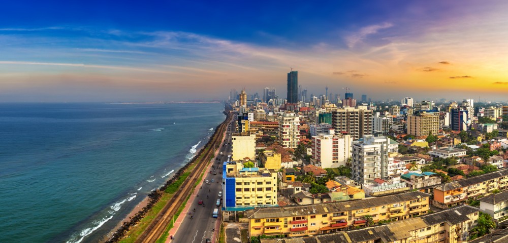 Coastal cityscape at sunset with high-rise buildings, a busy road, and the ocean stretching along the horizon.