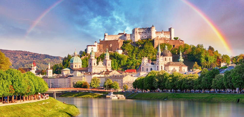 A vibrant rainbow arches over Salzburg, Austria, with the historic Hohensalzburg Fortress and baroque architecture reflecting in the calm river below.