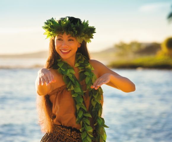 A woman dances joyfully near the ocean's edge, wearing a leaf crown and lei. The background features a serene shoreline with soft, golden light.