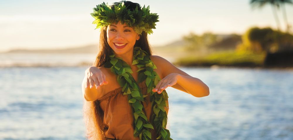 A woman dances joyfully near the ocean's edge, wearing a leaf crown and lei. The background features a serene shoreline with soft, golden light.