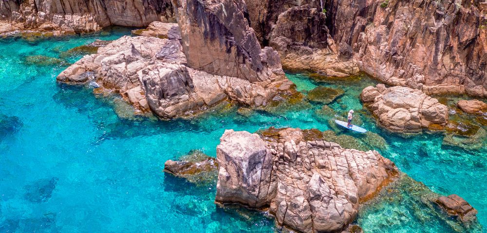 A person paddleboarding through clear turquoise waters surrounded by rugged, rocky cliffs. The serene scene conveys a sense of adventure and tranquility.