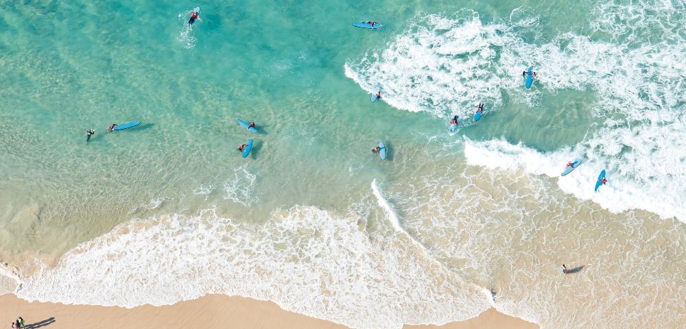 Aerial view of surfers on blue ocean waves near a sandy beach. Surfers on blue boards are waiting for waves, creating a lively, summery scene.
