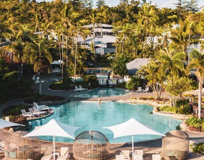 A serene resort pool surrounded by lush palm trees and greenery. Sun loungers and shaded areas offer relaxation. The scene conveys tropical tranquility.