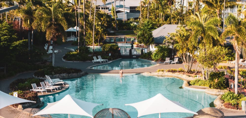 A serene resort pool surrounded by lush palm trees and greenery. Sun loungers and shaded areas offer relaxation. The scene conveys tropical tranquility.