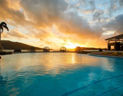 Sunset over a tranquil pool with palm trees and a distant mountain silhouette. The warm glow reflects on the water, creating a serene ambiance.