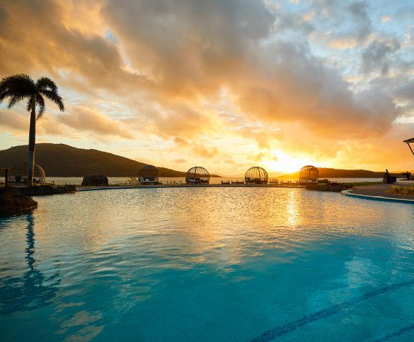 Sunset over a tranquil pool with palm trees and a distant mountain silhouette. The warm glow reflects on the water, creating a serene ambiance.