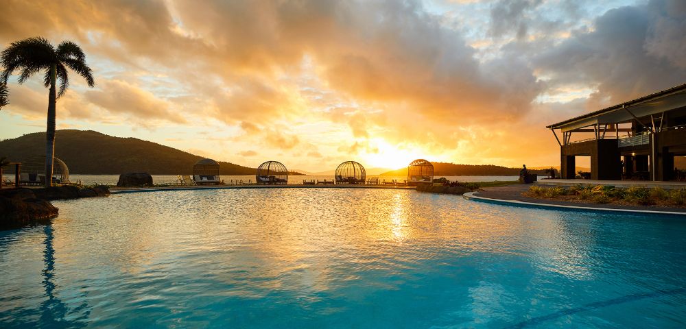 Sunset over a tranquil pool with palm trees and a distant mountain silhouette. The warm glow reflects on the water, creating a serene ambiance.