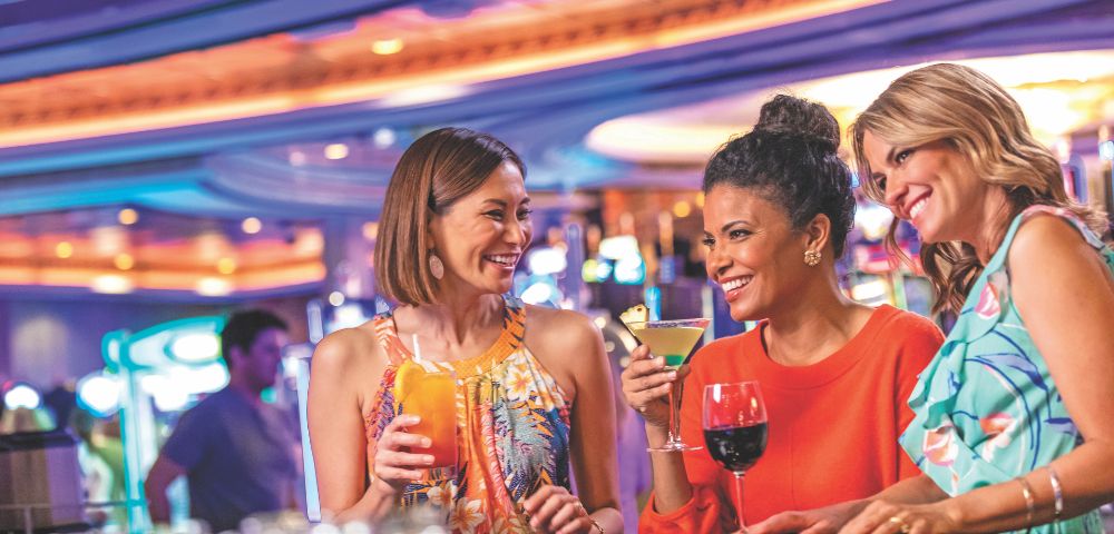Three women enjoy drinks together at a lively bar, smiling and chatting in a vibrant social atmosphere.